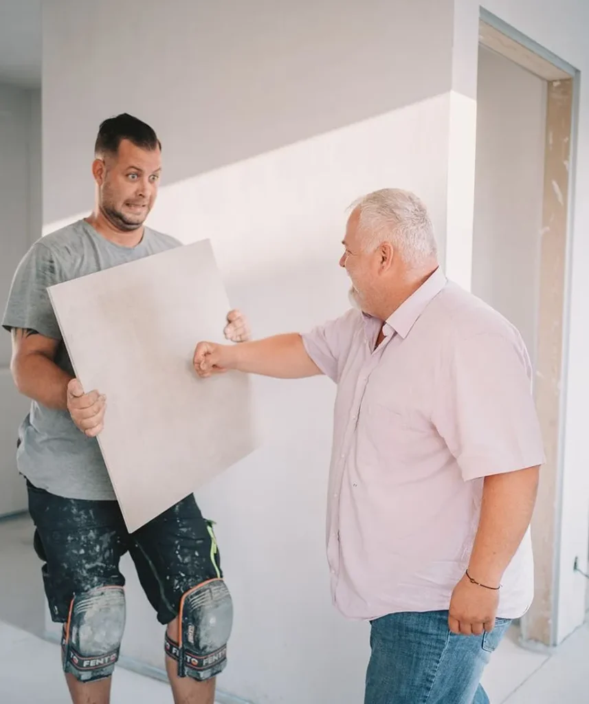 Fliesenleger hält Großformatfliese während einer humorvollen Baustellenszene